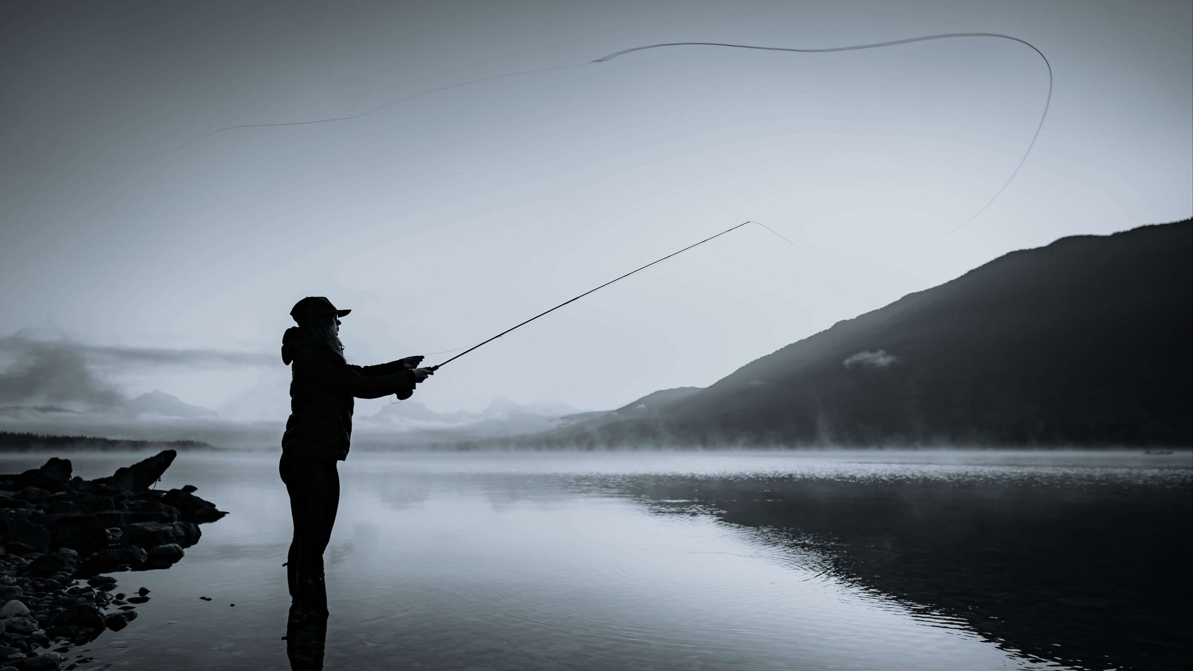 Silhouette of a person fly fishing on a misty lake with mountains in the background
